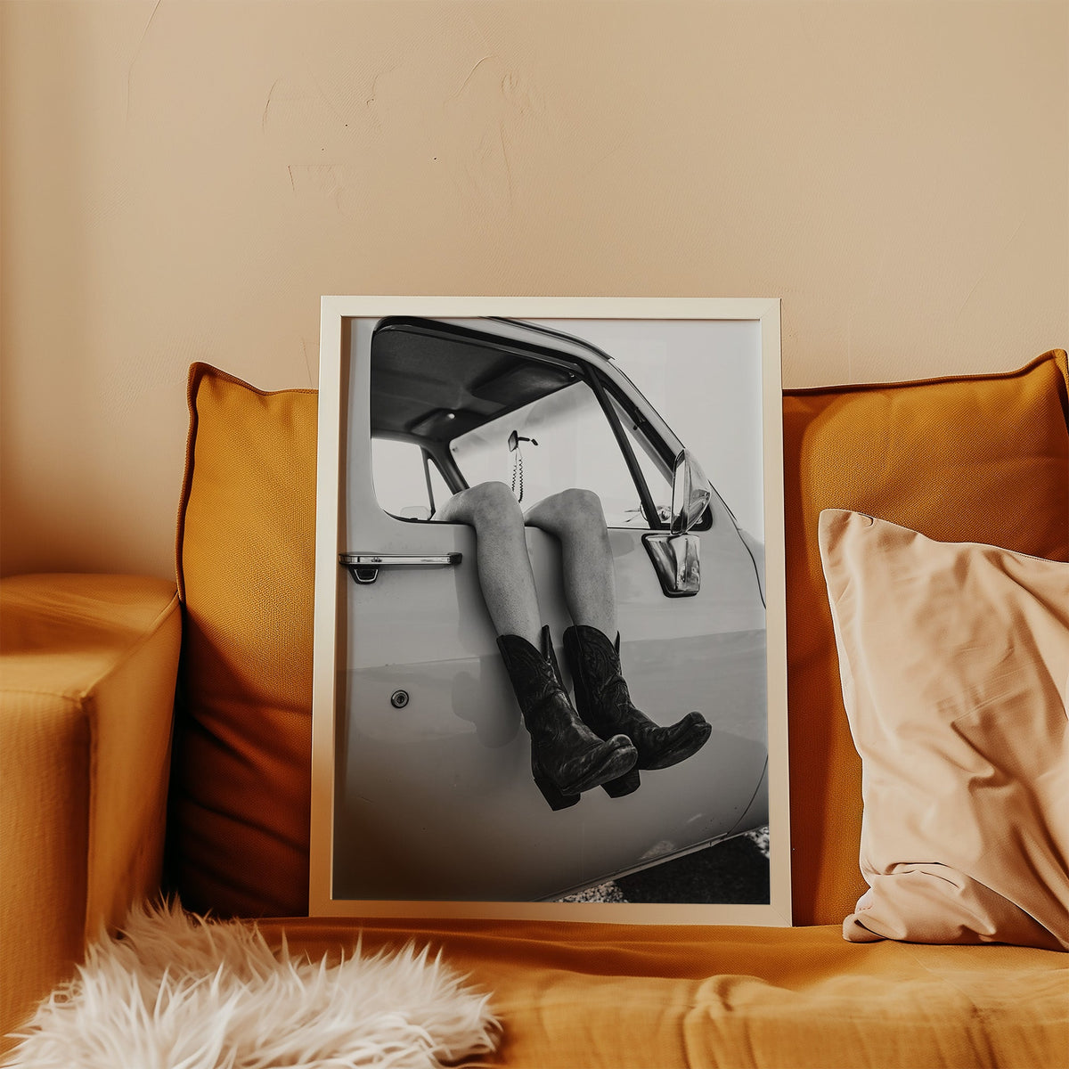 Cowboy boots dangling from a truck window print in a white frame leaning on a mustard velvet sofa