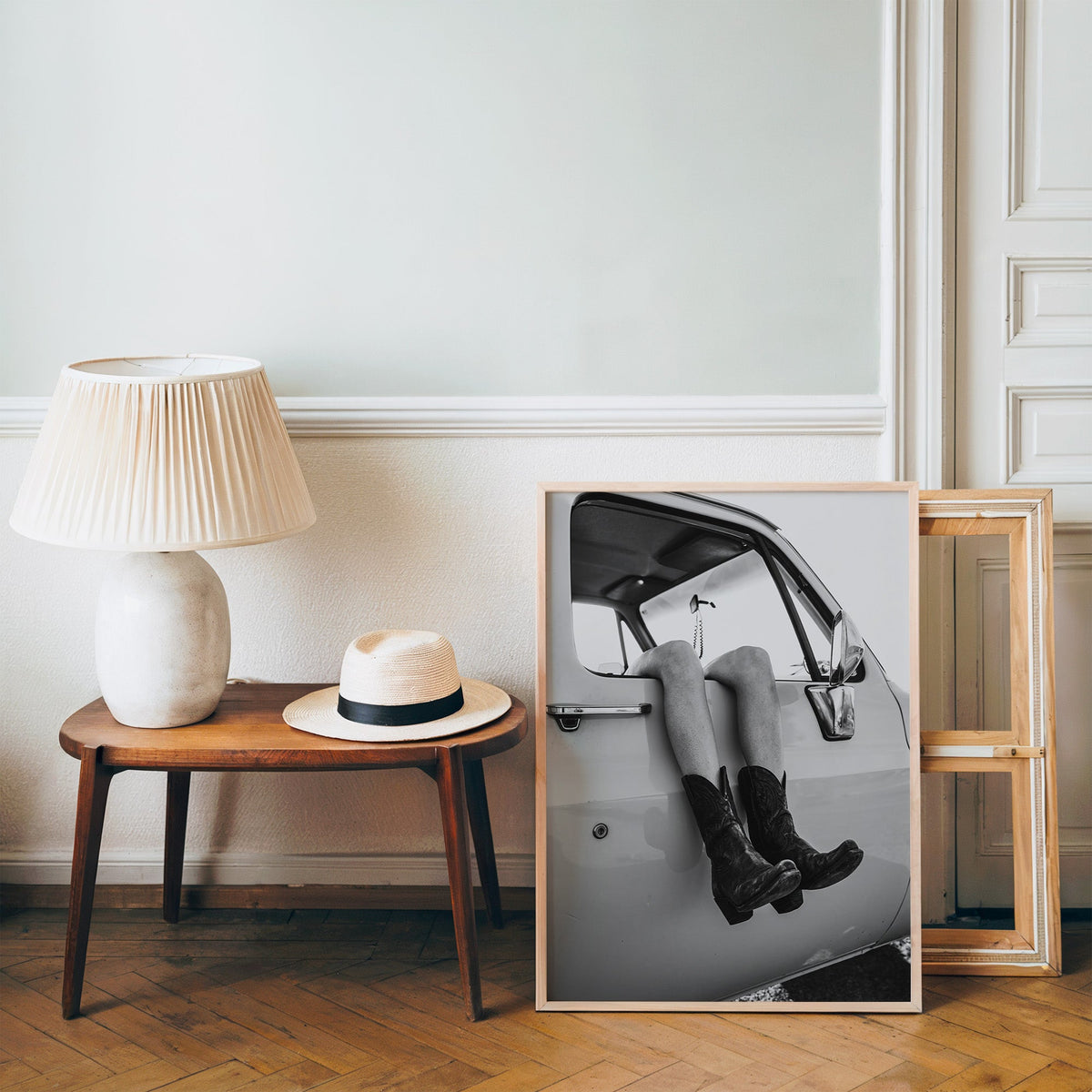 Cowboy boots dangling from a truck window print in a pine frame on a wood floor beside a lamp and straw hat