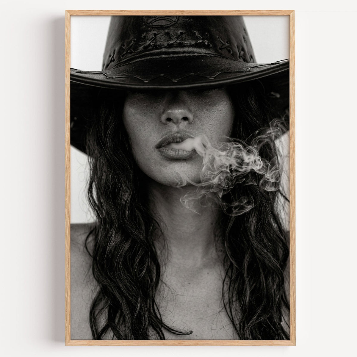 Cowboy hat brim and smoke from lips closeup print in a light oak frame on a plain white wall
