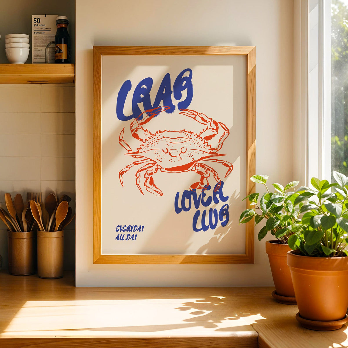 Crab Lover Club print in a light wood frame on a butcher block counter by crocks of wooden utensils