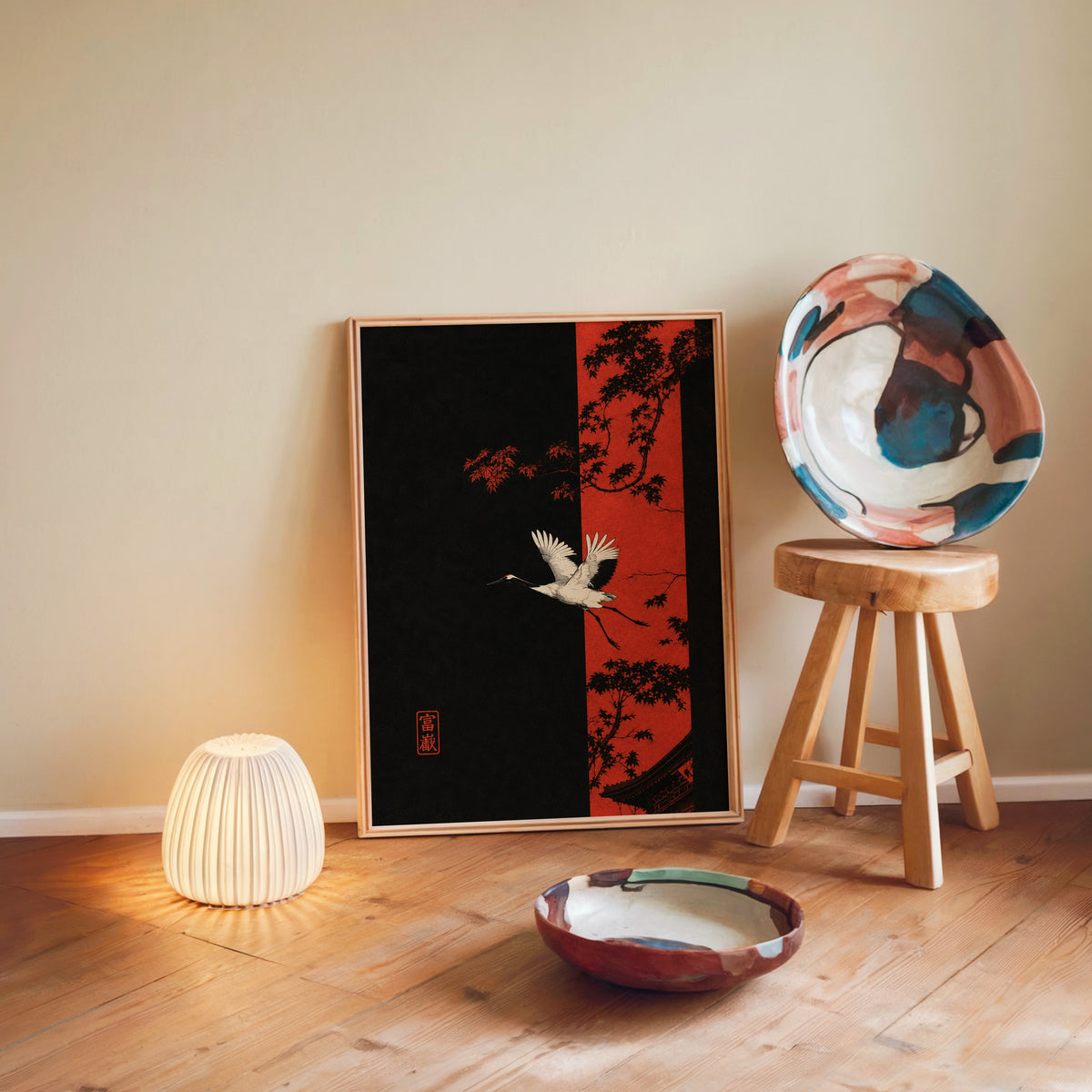white crane and red maple print in a light wood frame leaning on a wooden floor by a glowing lamp and stool