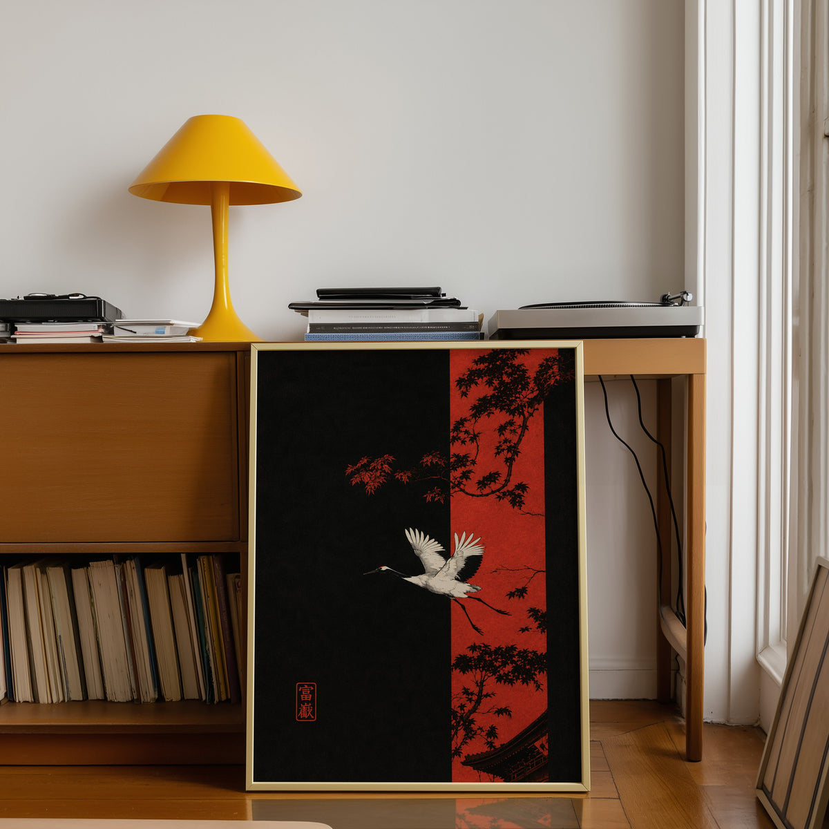 white crane and red maple print shown in a gold frame leaning under a teak desk with a yellow lamp
