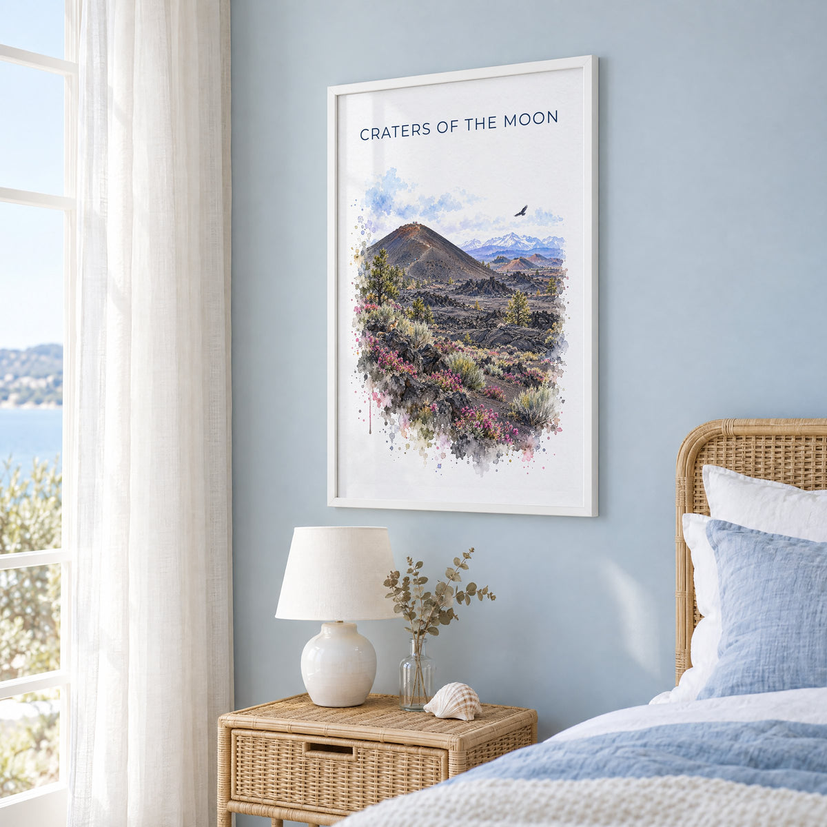 Craters of the Moon National Monument watercolor in a white frame on a pale blue wall beside a rattan bed in a coastal