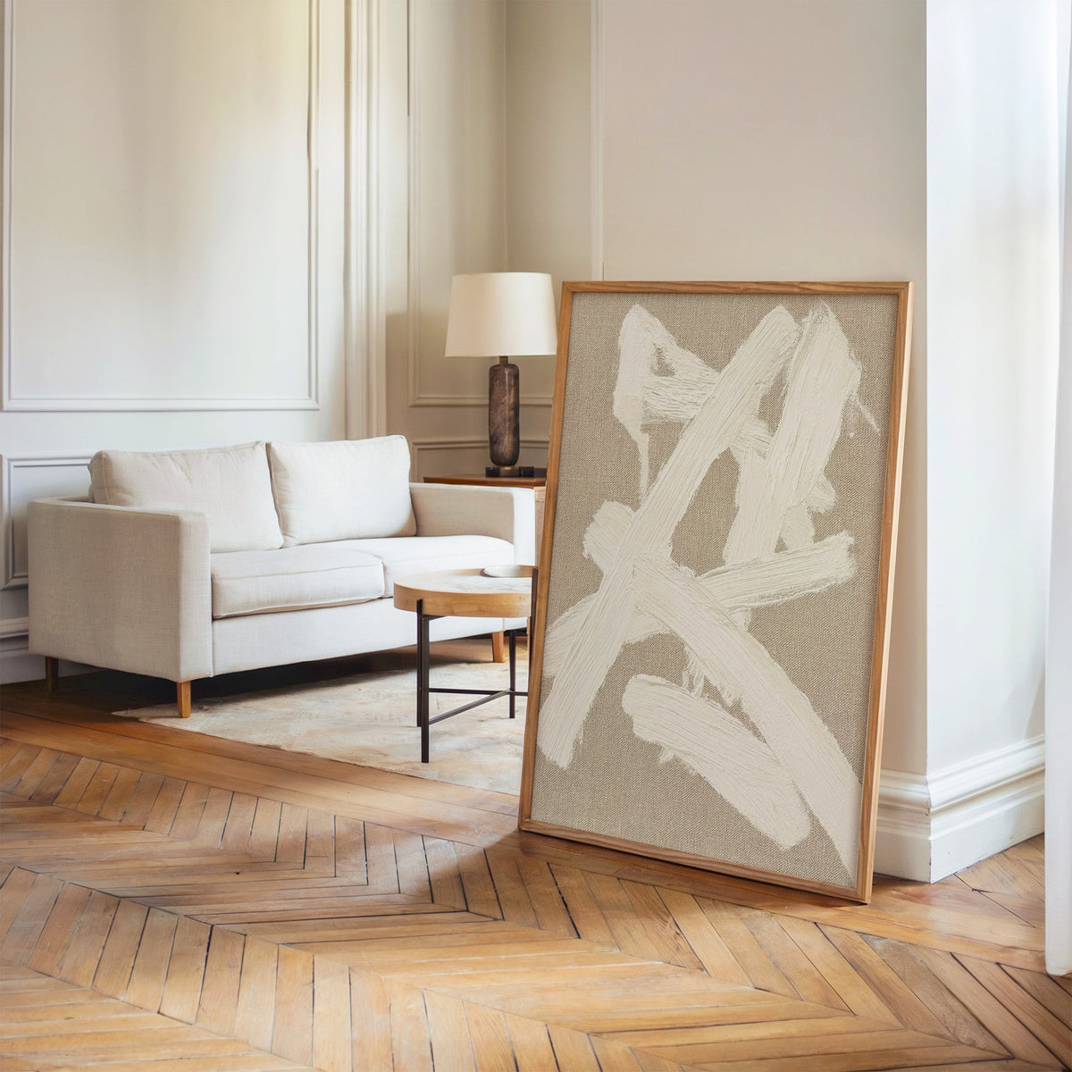 Cream impasto crossed strokes on linen print in an oak frame leaning on a herringbone floor by a cream linen sofa