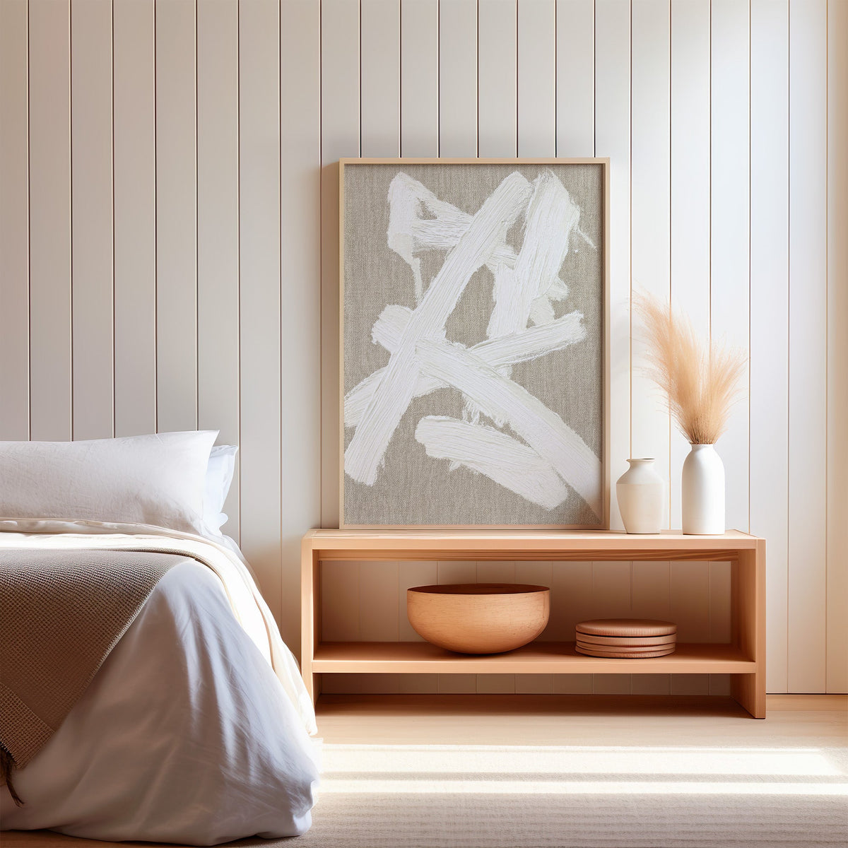 Cream impasto crossed strokes on linen print in a light oak frame on a wood bench by a bed with pampas grass