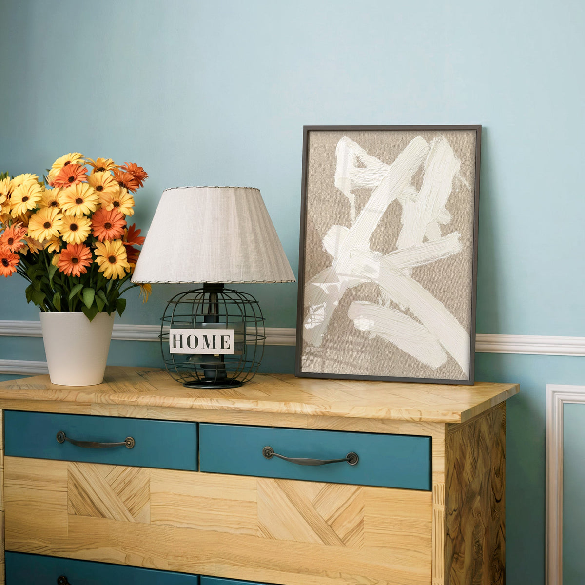 Cream impasto crossed strokes on linen print in a charcoal frame on a teal dresser beside orange and yellow daisies