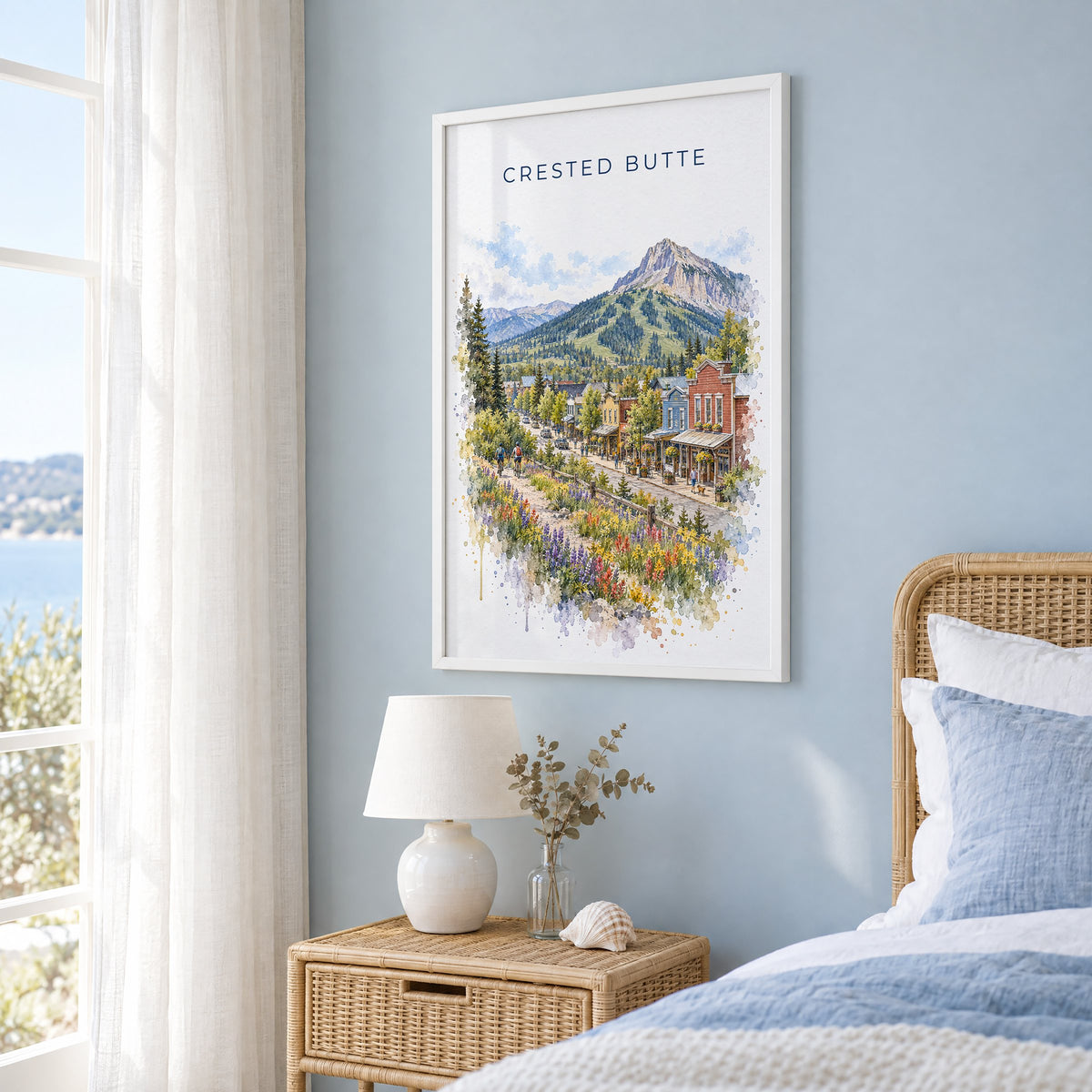 Crested Butte watercolor print in a white frame on a pale blue wall beside a rattan bed in a coastal bedroom