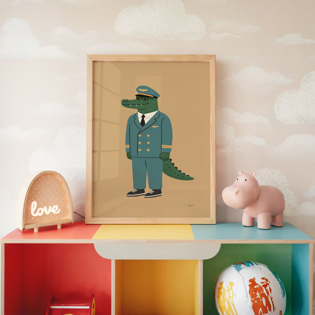 Crocodile in teal pilot uniform nursery print in an oak frame on a rainbow toy shelf against cloud wallpaper