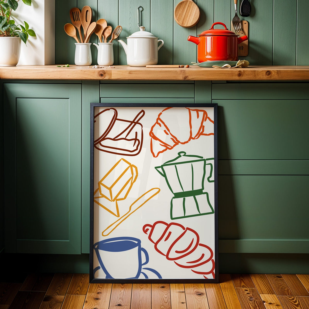 Croissant and coffee doodle print in a black frame against sage green kitchen cabinets