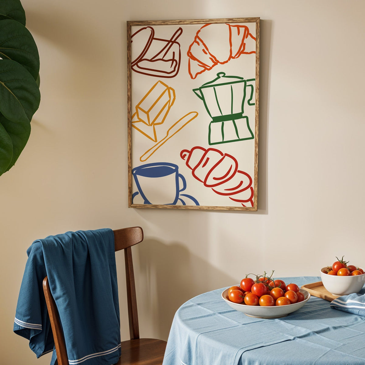 Croissant and coffee doodle print in an oak frame above a blue linen table with ripe tomatoes