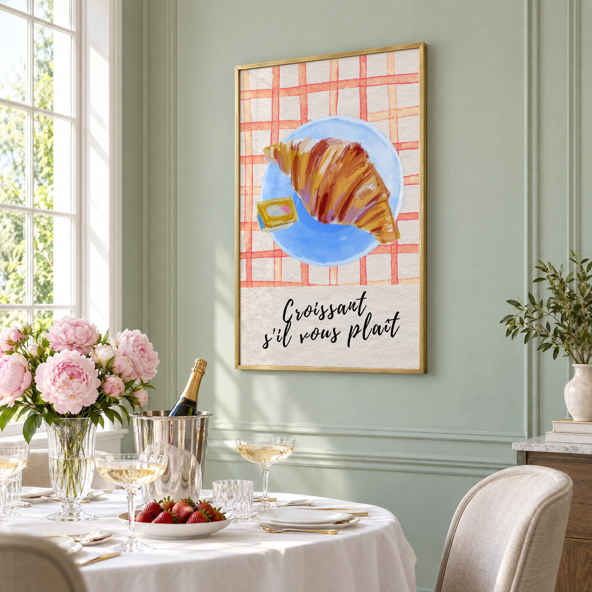 Croissant s'il vous plait blue plate print in a brass frame on a sage wall above a brunch table with champagne and peonies, seen at an angle