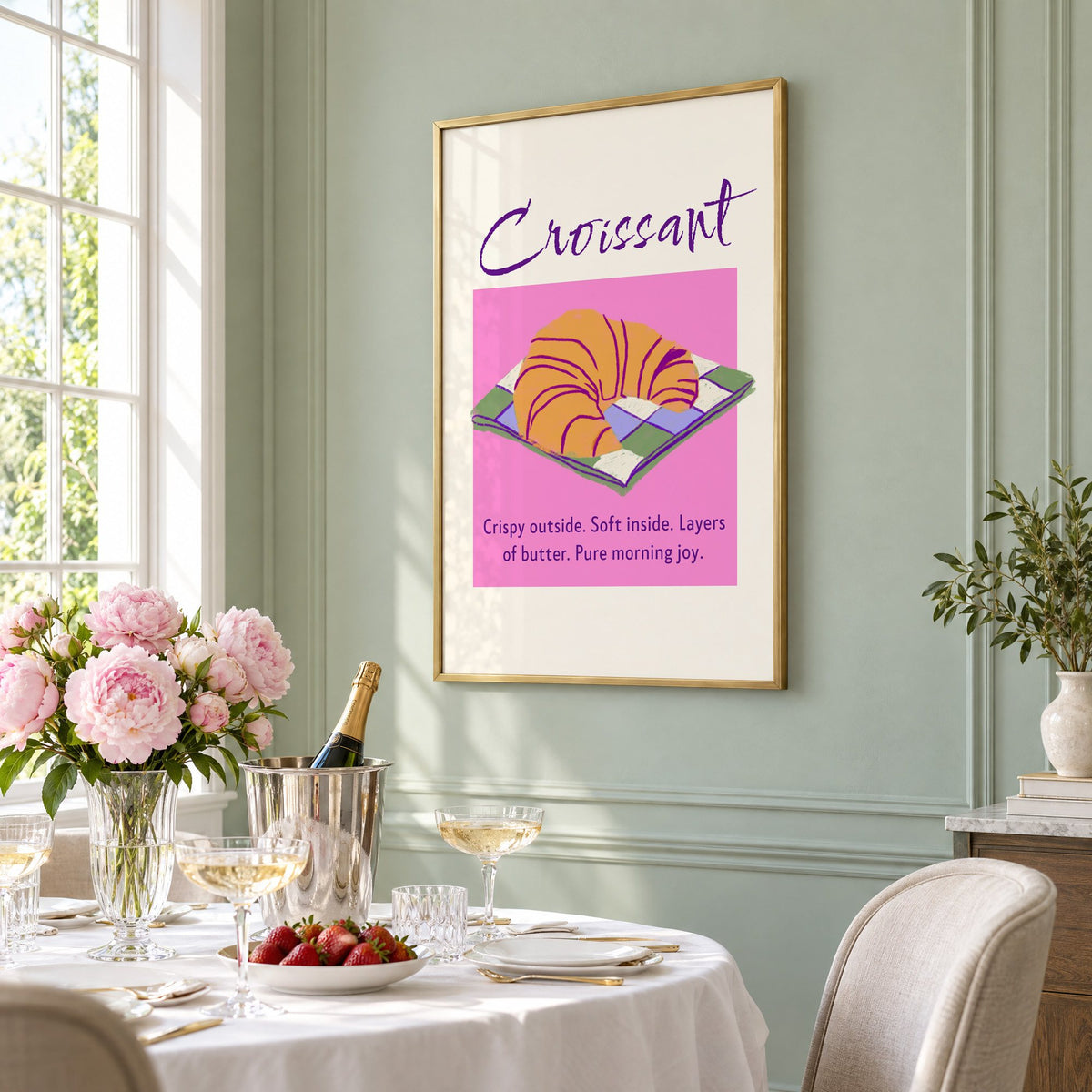 Croissant on checkered napkin pink panel print in a brass frame on a sage wall above a brunch table with champagne and peonies, seen at an angle