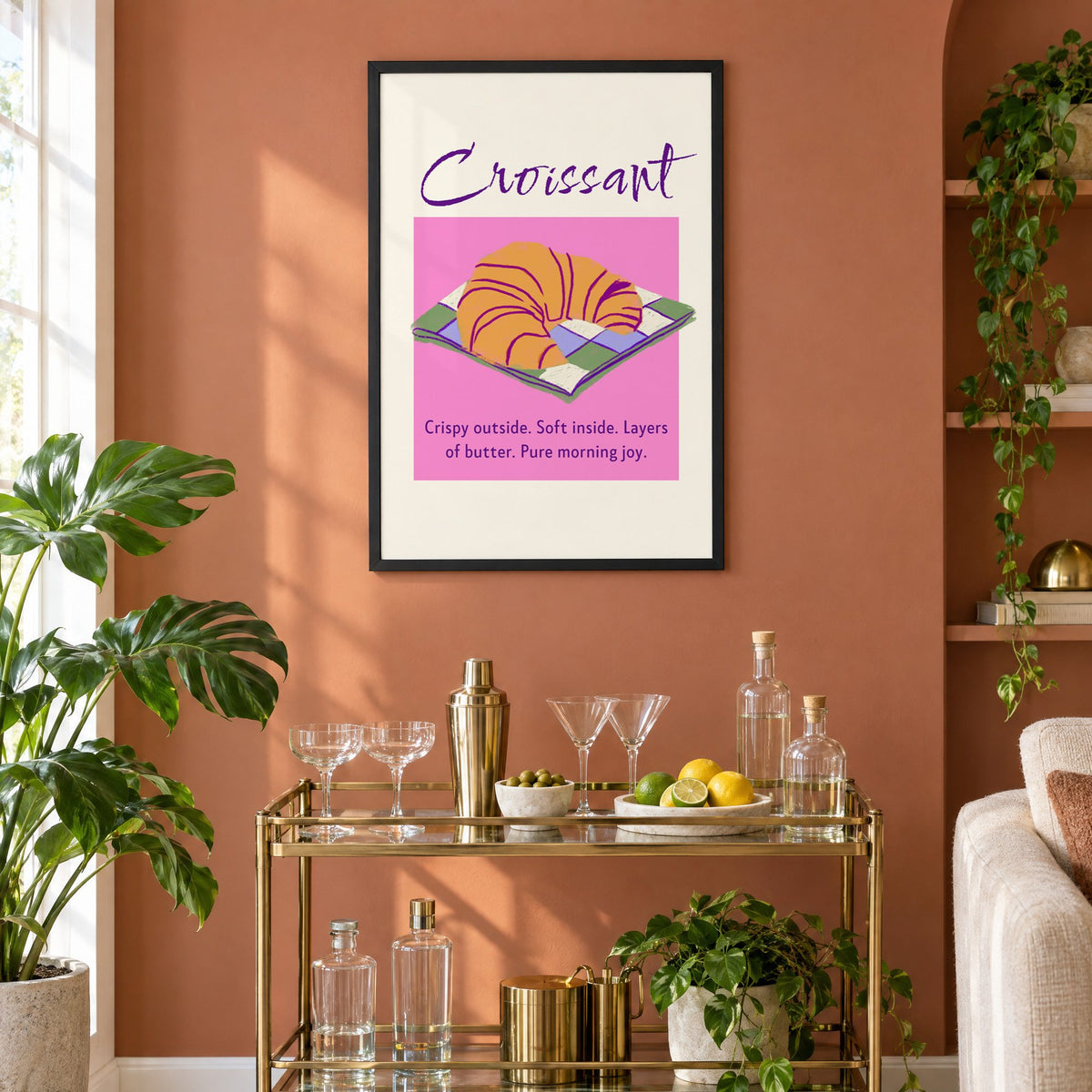 Croissant on checkered napkin pink panel print in a black frame on a terracotta wall above a brass bar cart with coupes and citrus