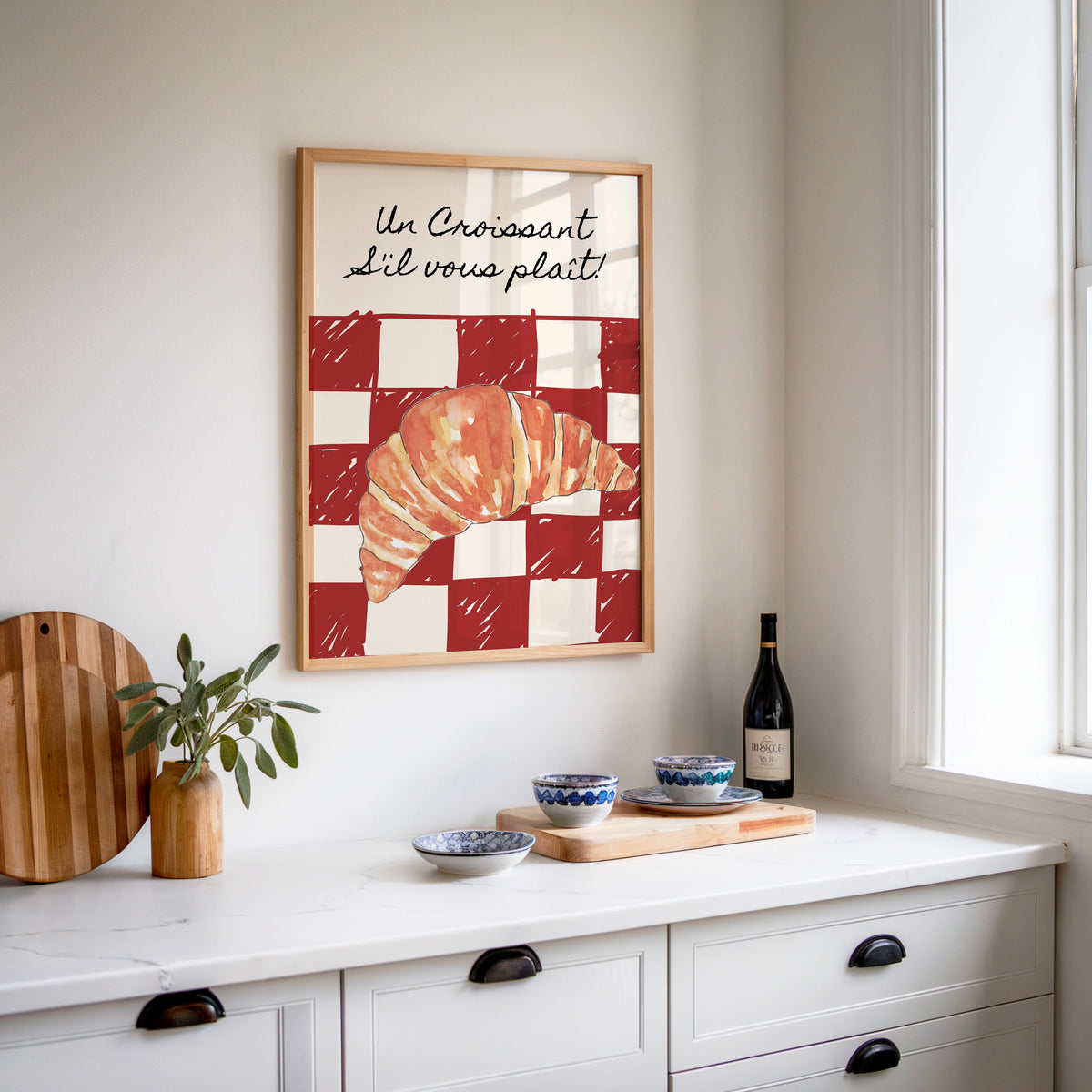 Un Croissant red check print in an oak frame above a white kitchen sideboard with a wine bottle and wooden board