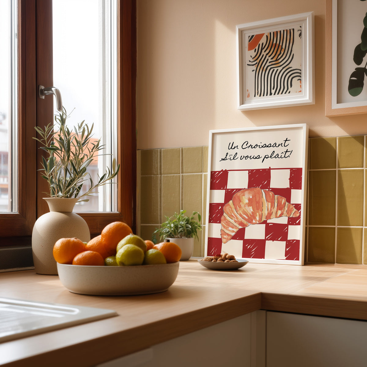 Un Croissant red check print in a white frame leaning against olive green kitchen tiles beside a bowl of citrus