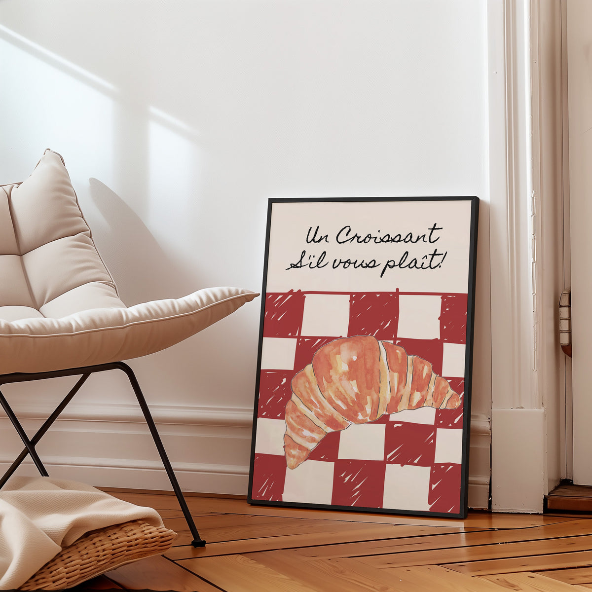 Un Croissant red check print in a slim black frame leaning on a herringbone floor beside a cream armchair