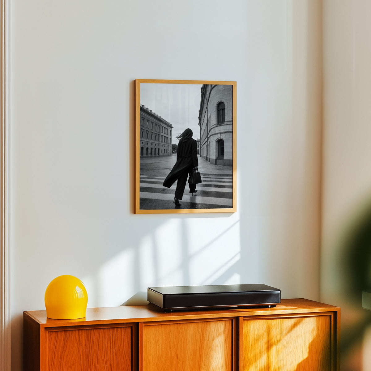Woman in long black coat on wet crosswalk print in an oak frame above a teak sideboard with a yellow glass lamp