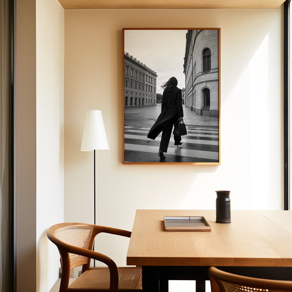 Woman in long black coat on wet crosswalk print in an oak frame above a dining table and cane chair