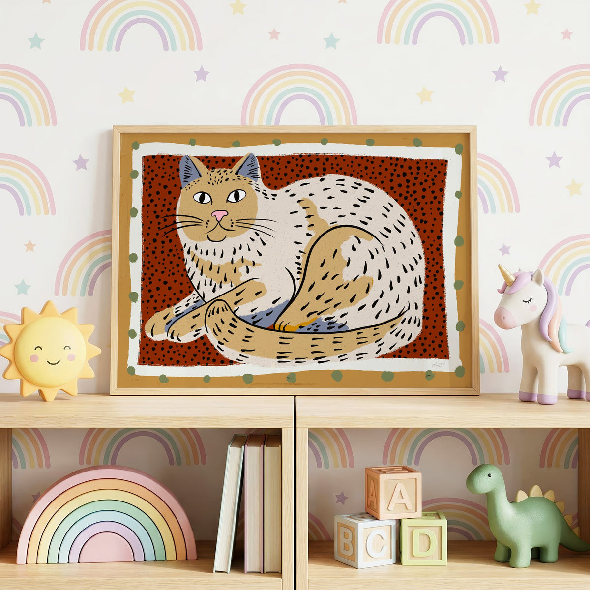 Curled cream cat on rust dots nursery print in an oak frame on a toy shelf against rainbow wallpaper
