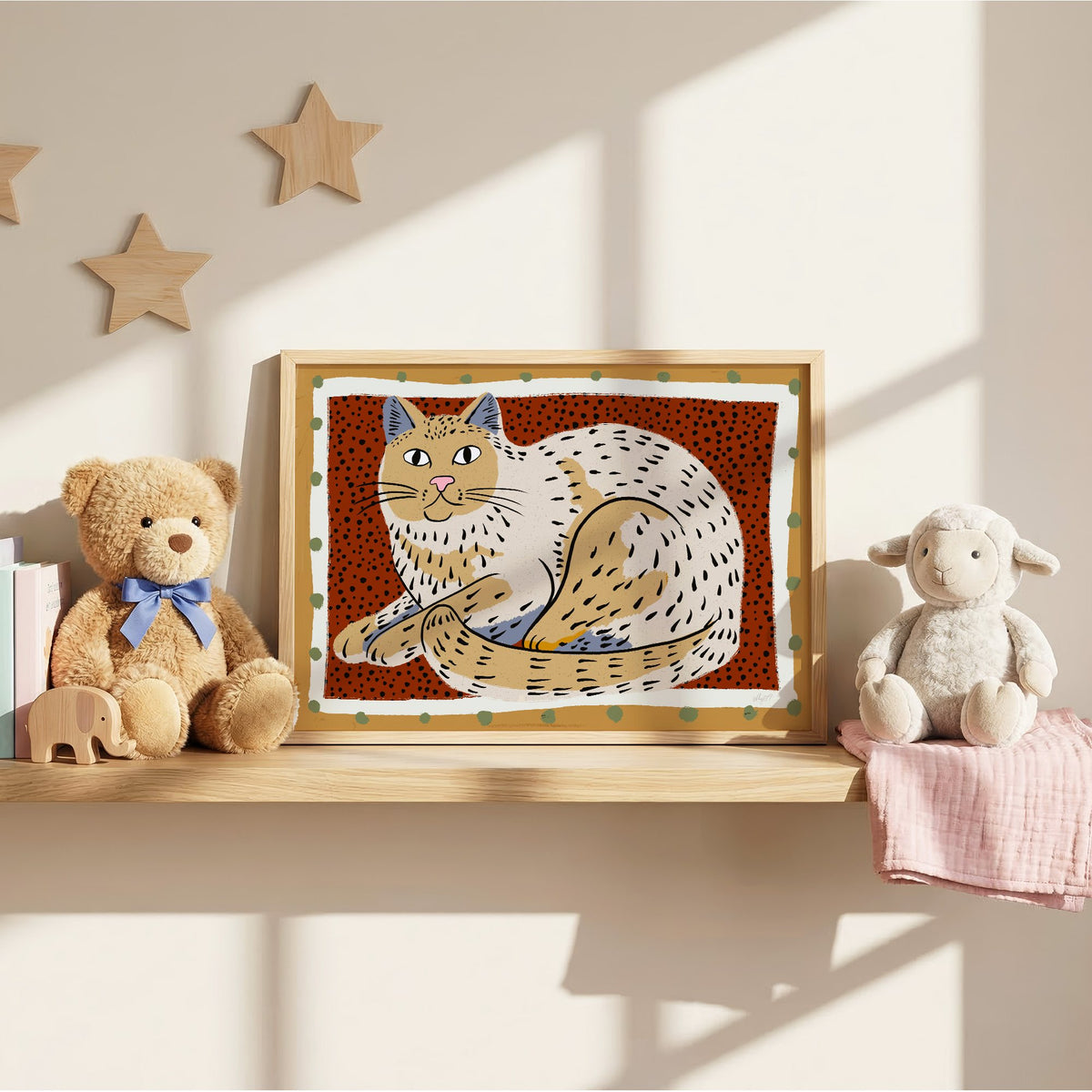Curled cream cat on rust dots nursery print in an oak frame on a wall shelf between a teddy bear and a toy lamb