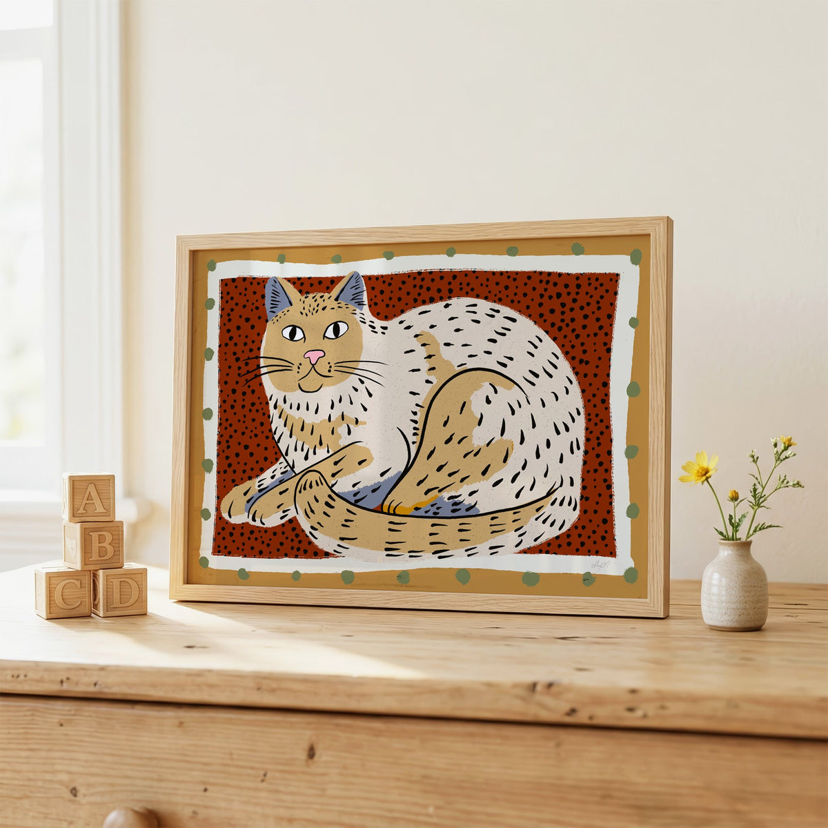 Curled cream cat on rust dots nursery print in an oak frame on a wooden dresser beside ABC blocks