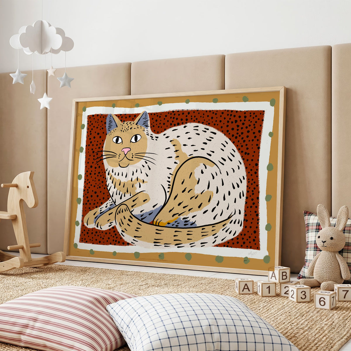 Curled cream cat on rust dots nursery print in an oak frame on a padded wall by a wooden rocking horse
