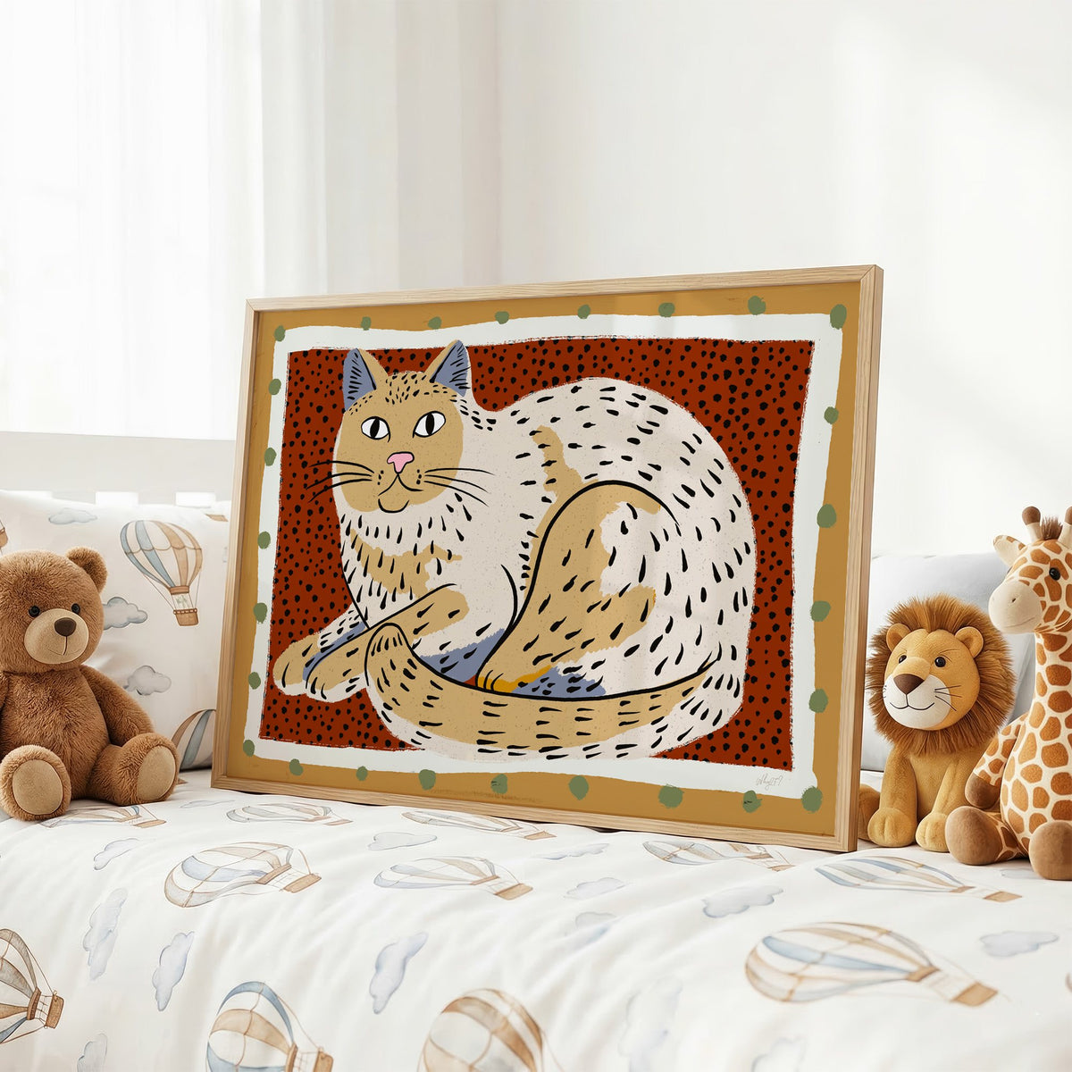 Curled cream cat on rust dots nursery print in an oak frame on a kids bed with hot air balloon bedding