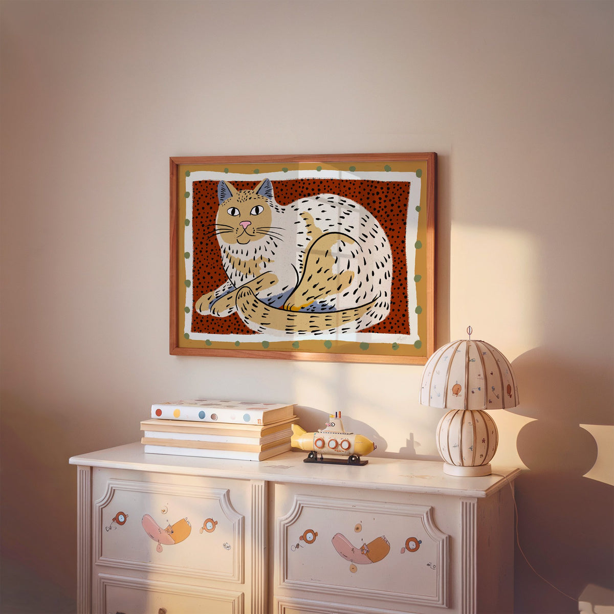 Curled cream cat on rust dots nursery print in a cherry wood frame above a painted dresser and lamp