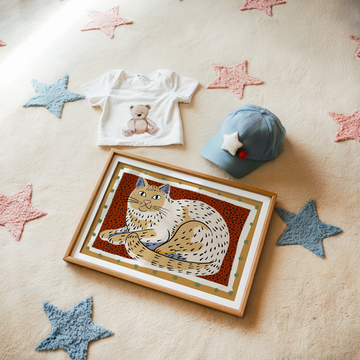 Curled cream cat on rust dots nursery print in a matted oak frame on a star rug with a baby tee