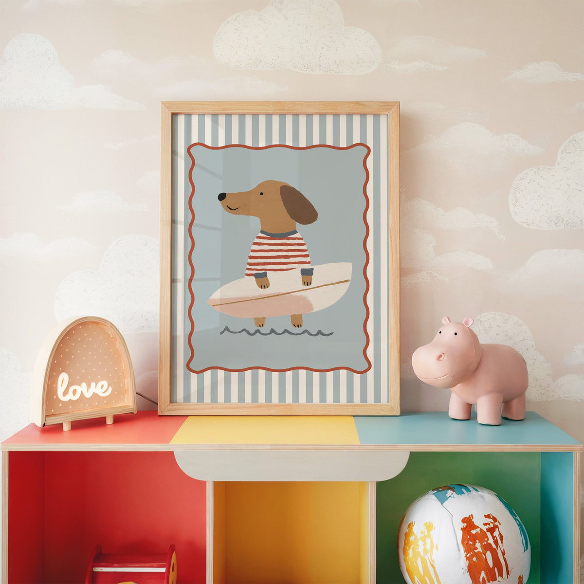 Dachshund holding surfboard nursery print in an oak frame on a rainbow toy shelf against cloud wallpaper