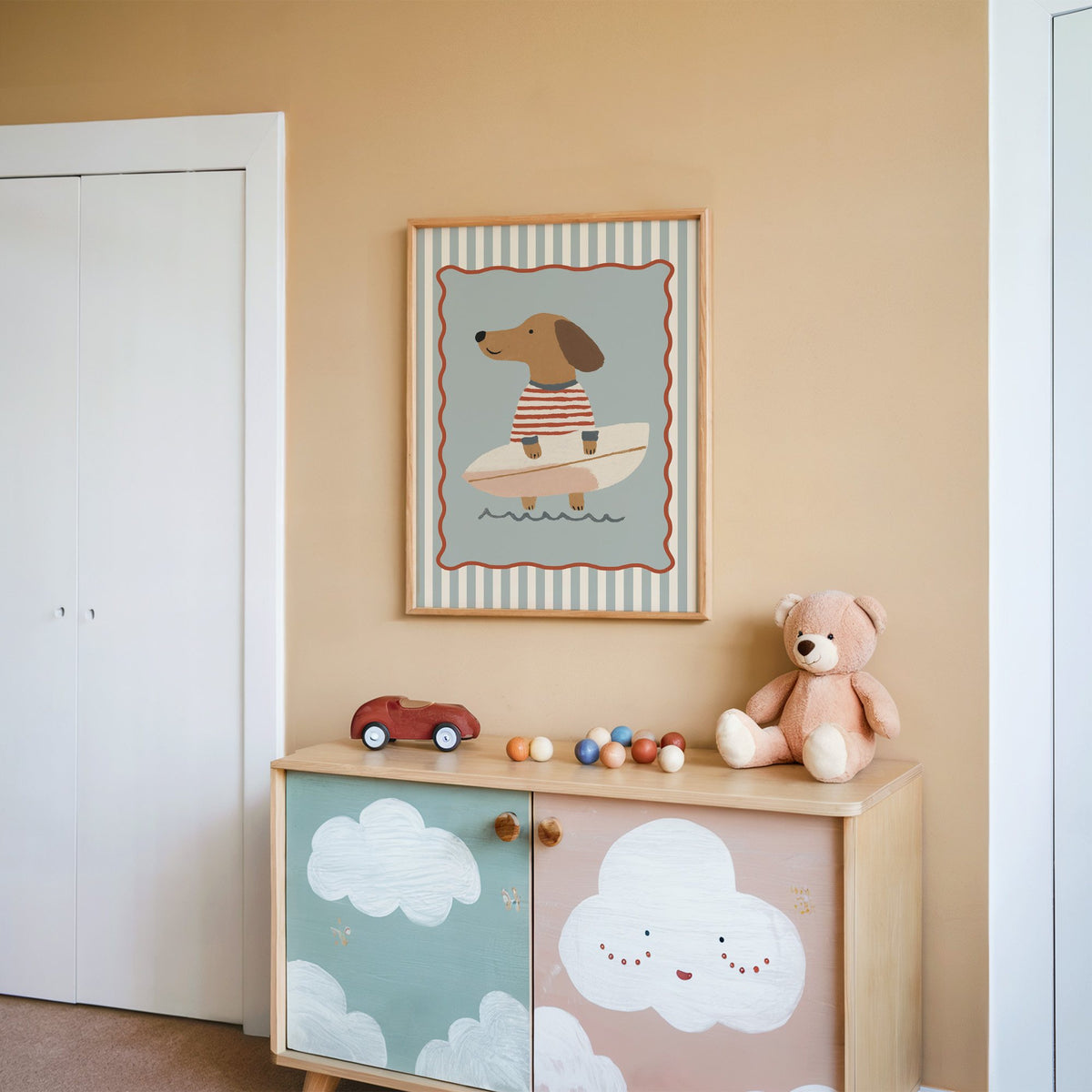 Dachshund holding surfboard nursery print in an oak frame on a peach wall above a cloud-painted cabinet