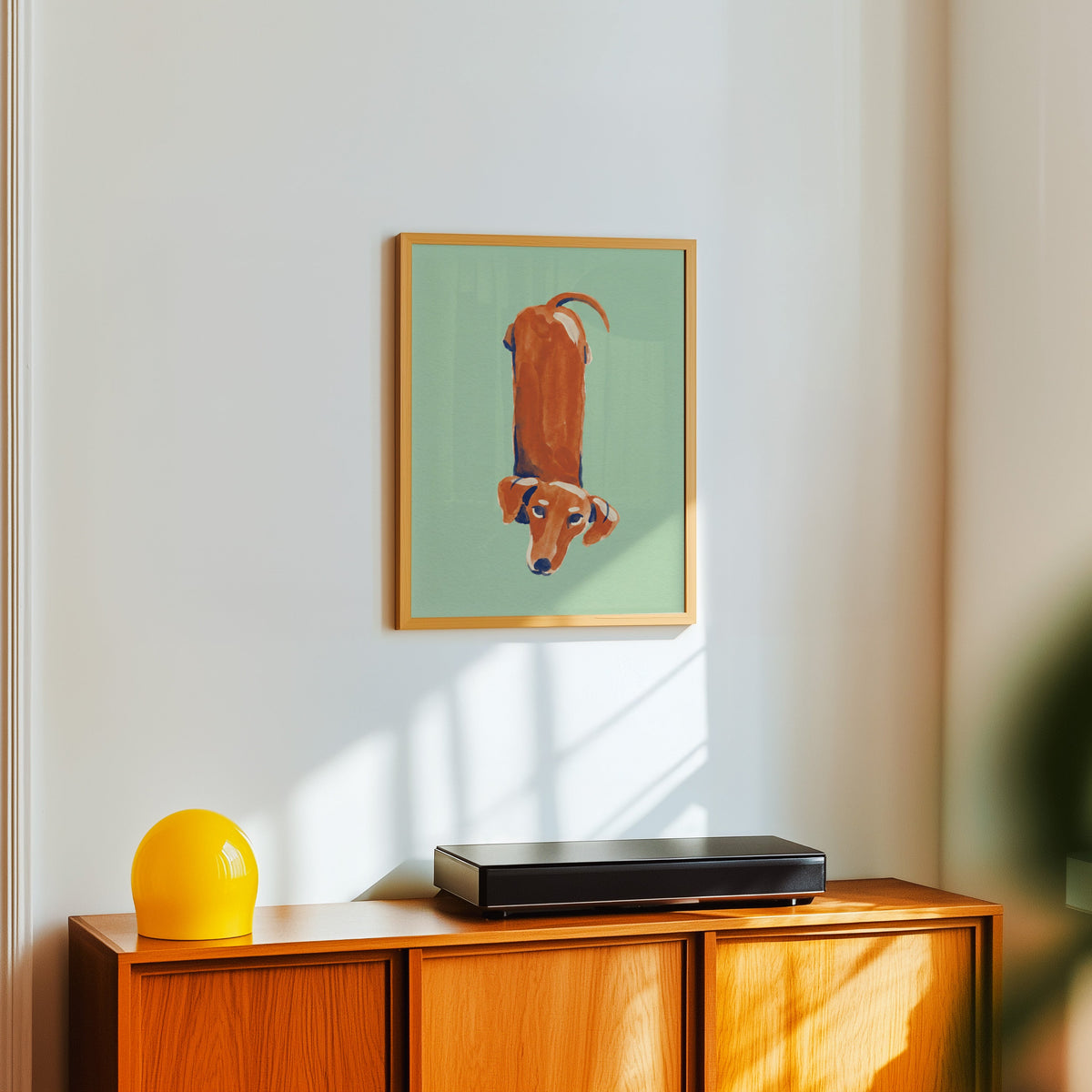 Funny dachshund print in a light oak frame above a teak sideboard with a yellow lamp