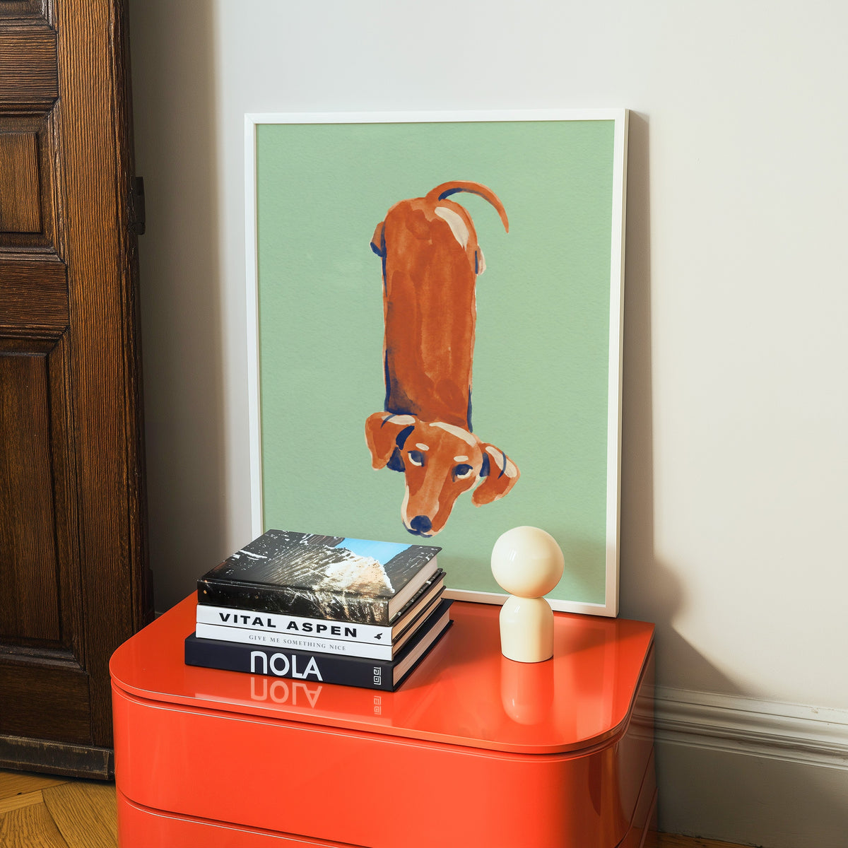 Sausage dog print in a white frame on a glossy orange cabinet with books