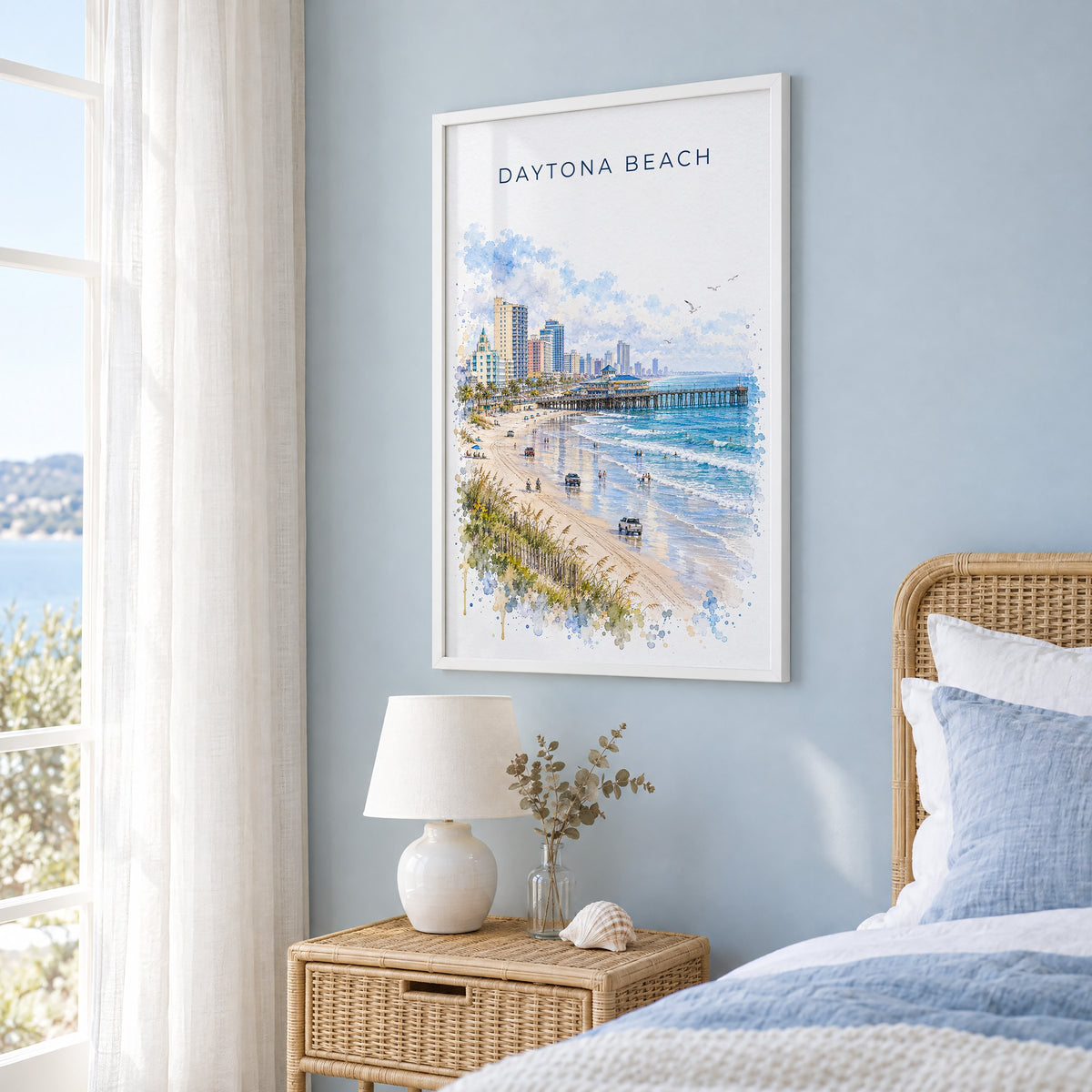 Daytona Beach watercolor print in a white frame on a pale blue wall beside a rattan bed in a coastal bedroom