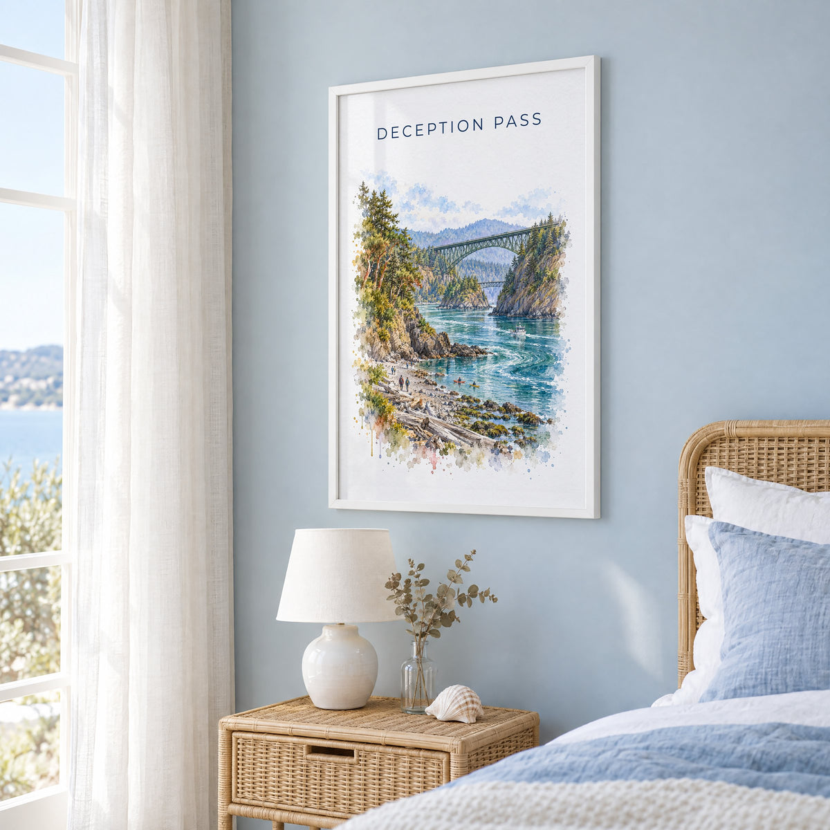Deception Pass watercolor print in a white frame on a pale blue wall beside a rattan bed in a coastal bedroom