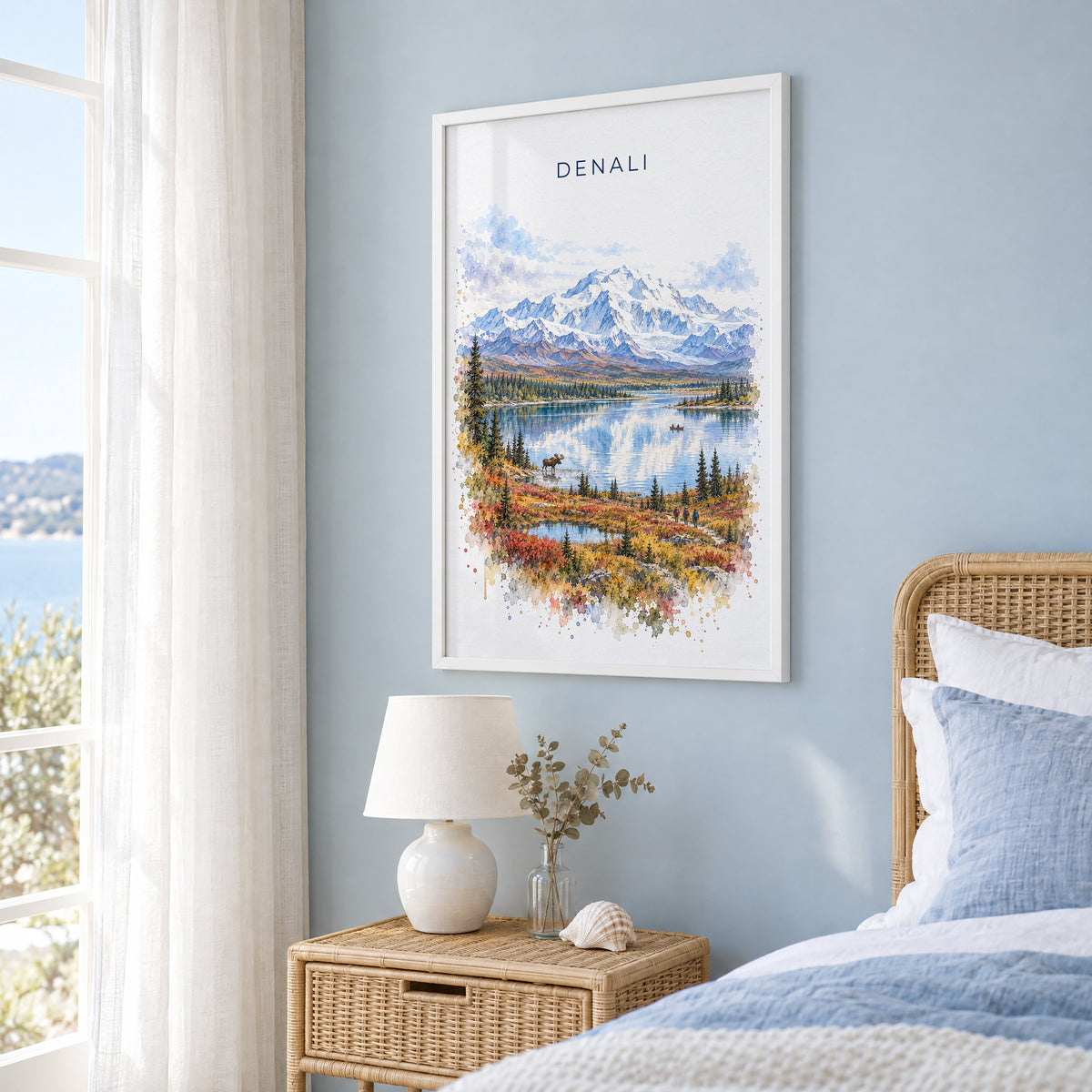 Denali National Park watercolor print in a white frame on a pale blue wall beside a rattan bed in a coastal bedroom