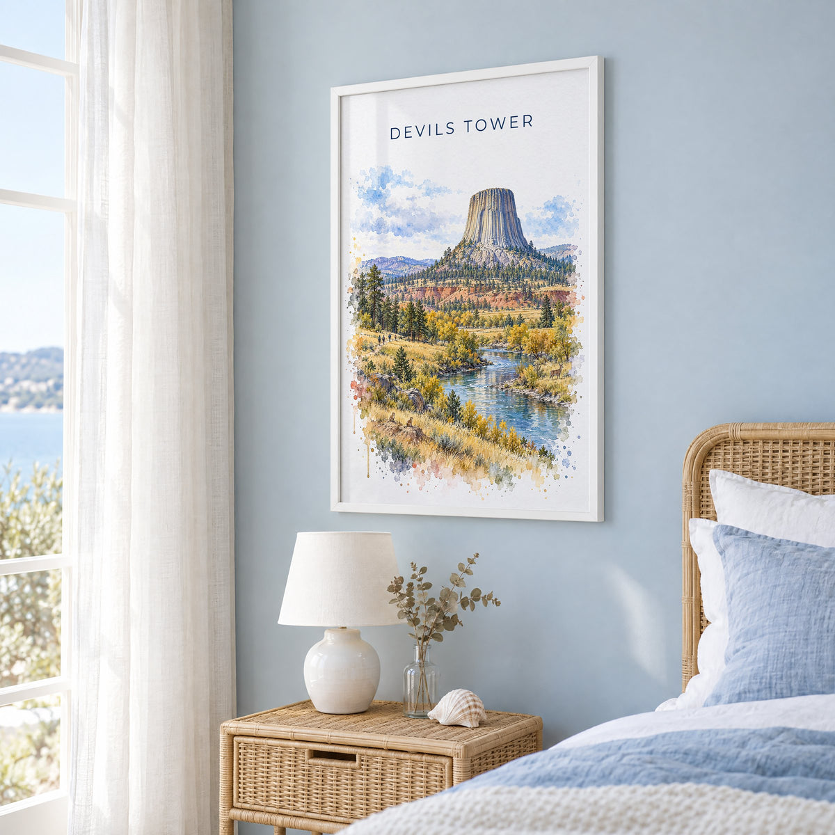 Devils Tower watercolor print in a white frame on a pale blue wall beside a rattan bed in a coastal bedroom