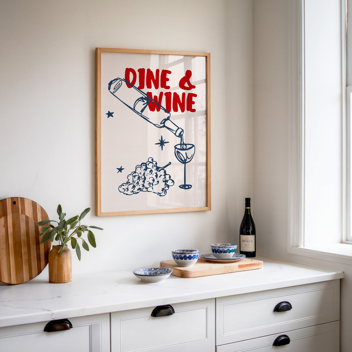 Dine and Wine print in an oak frame above a white sideboard with a bottle of wine