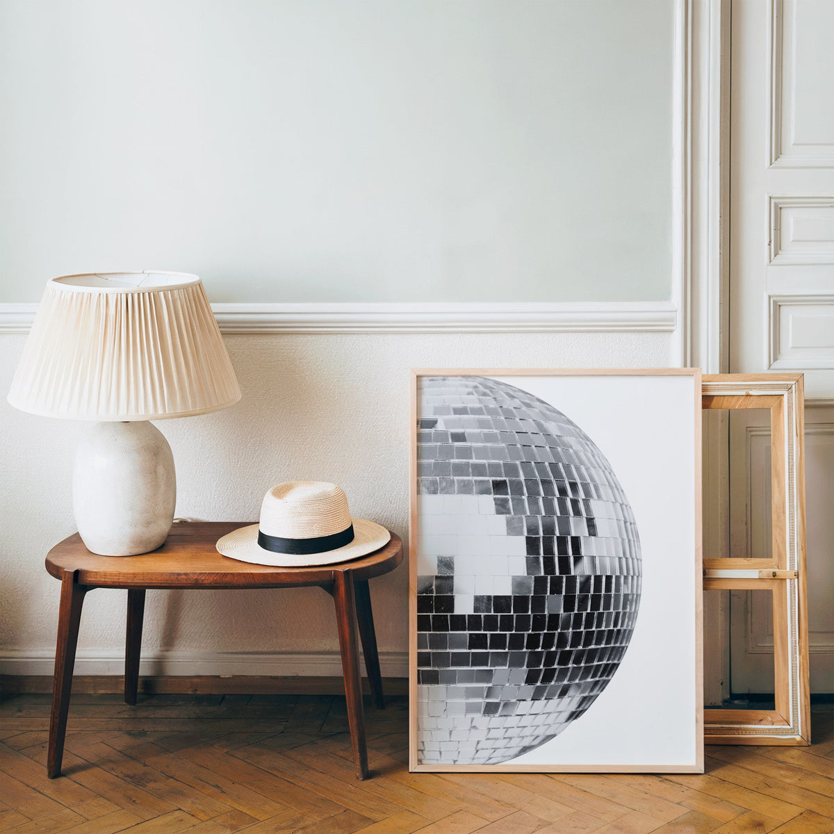 Disco mirror ball close-up print in a pine frame on a wood floor beside a lamp and straw hat