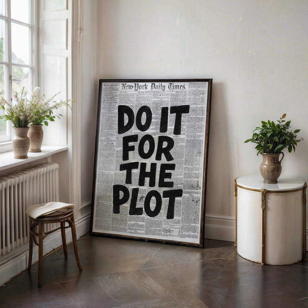 Do It for the Plot black lettering on old newspaper print in a black frame on a dark stone floor beside a white side table