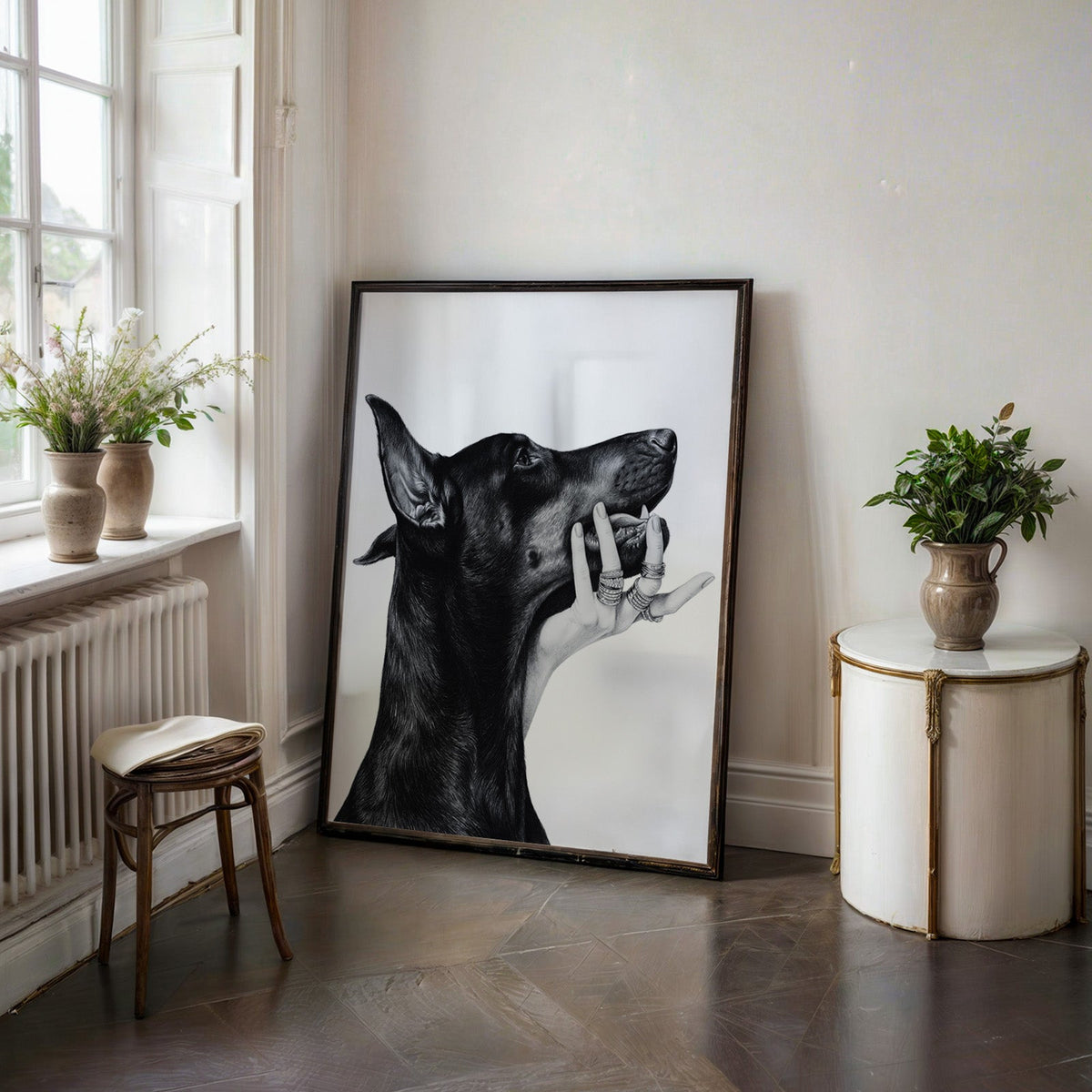 Drawn Doberman with ring-covered hand under chin print in a black frame on a dark stone floor beside a white side table