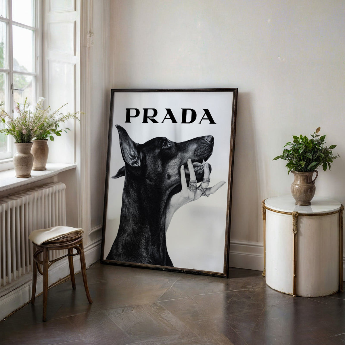 Drawn Doberman with ringed hand under chin print in a black frame on a dark stone floor beside a white side table