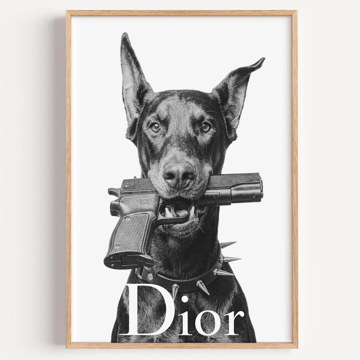 Doberman holding toy pistol with white lettering print in a light oak frame on a plain white wall