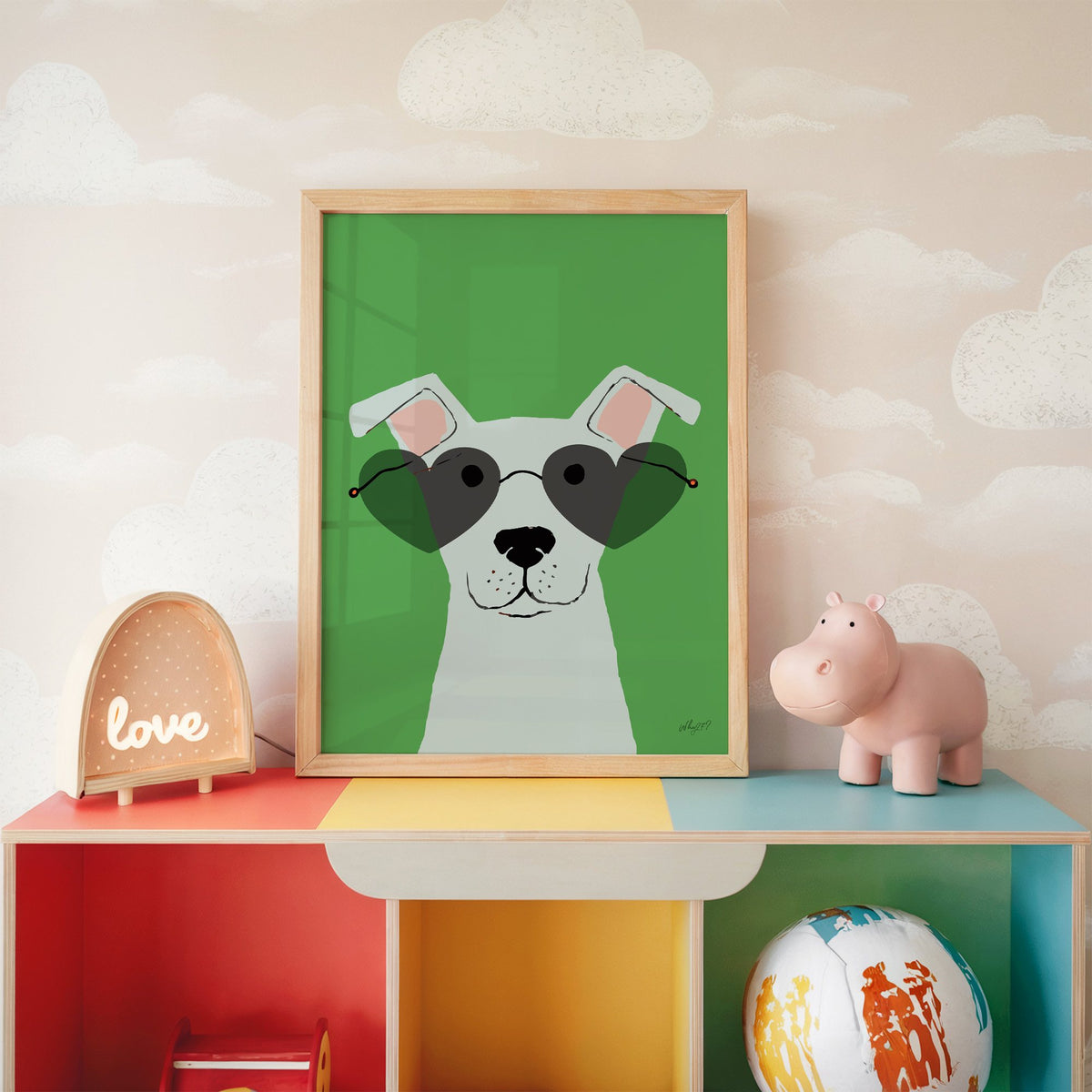 White dog in heart sunglasses nursery print in an oak frame on a rainbow toy shelf against cloud wallpaper