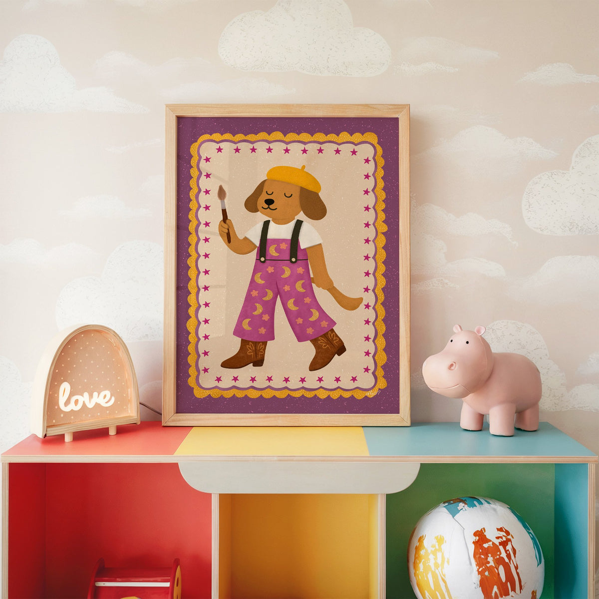 Painter dog in yellow beret nursery print in an oak frame on a rainbow toy shelf against cloud wallpaper
