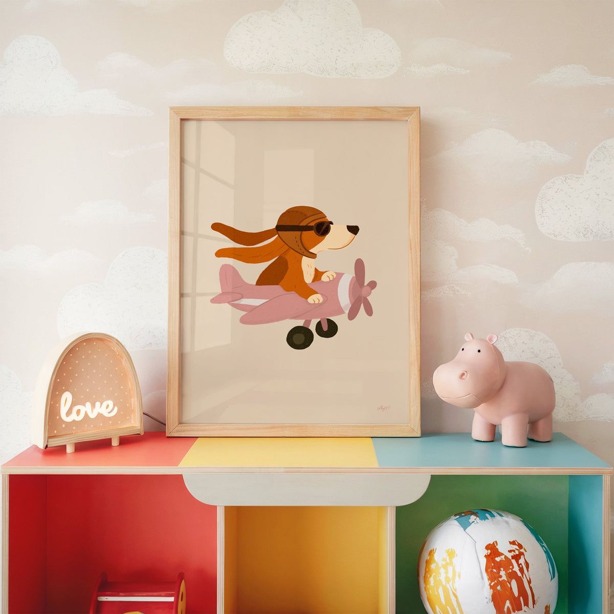 Dog pilot flying pink plane nursery print in an oak frame on a rainbow toy shelf against cloud wallpaper