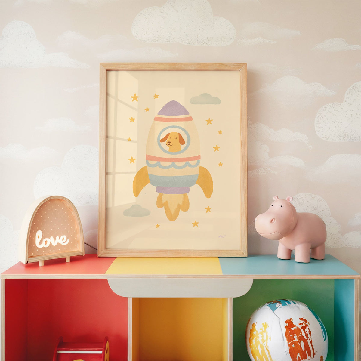 Smiling dog in pastel rocket nursery print in an oak frame on a rainbow toy shelf against cloud wallpaper
