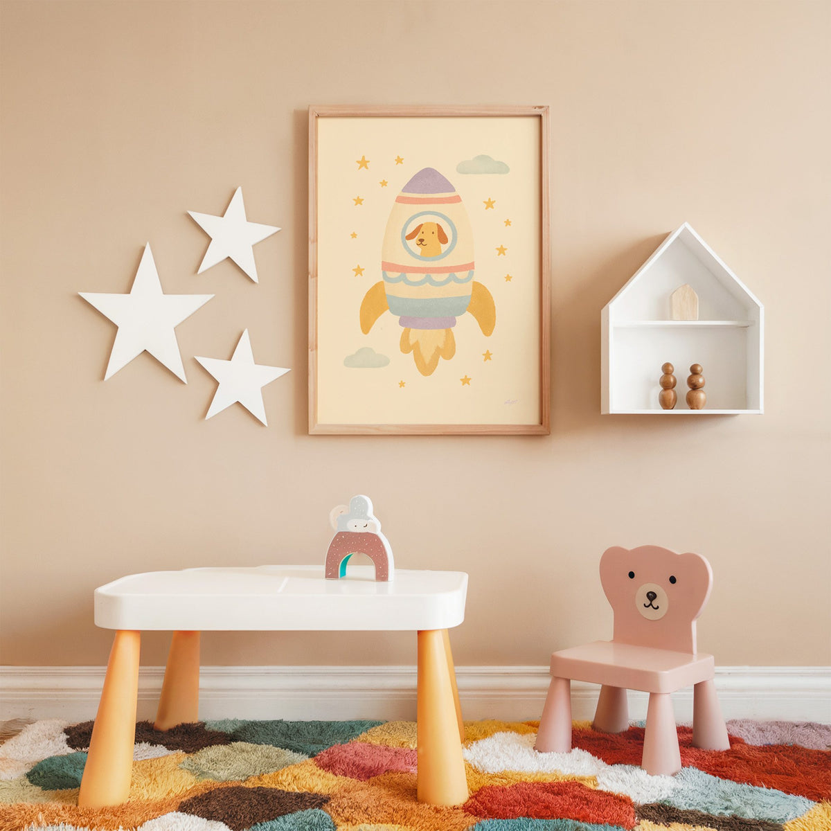 Smiling dog in pastel rocket nursery print in an oak frame on a peach wall above a kids table with wall stars