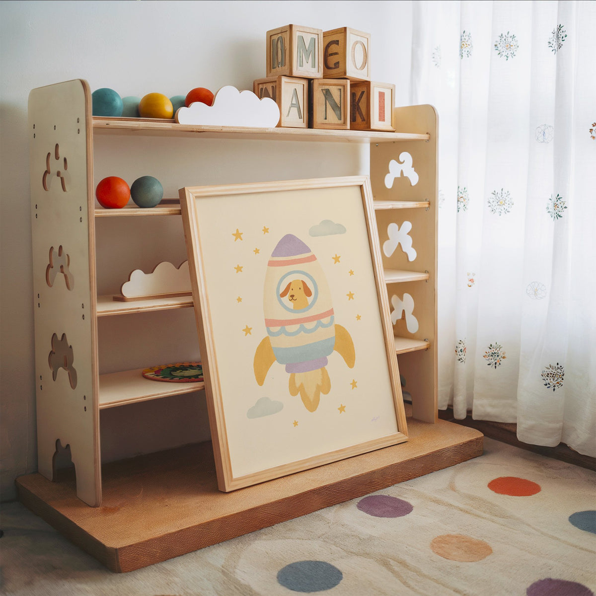 Smiling dog in pastel rocket nursery print in an oak frame leaning on a toy shelf with wooden letter blocks