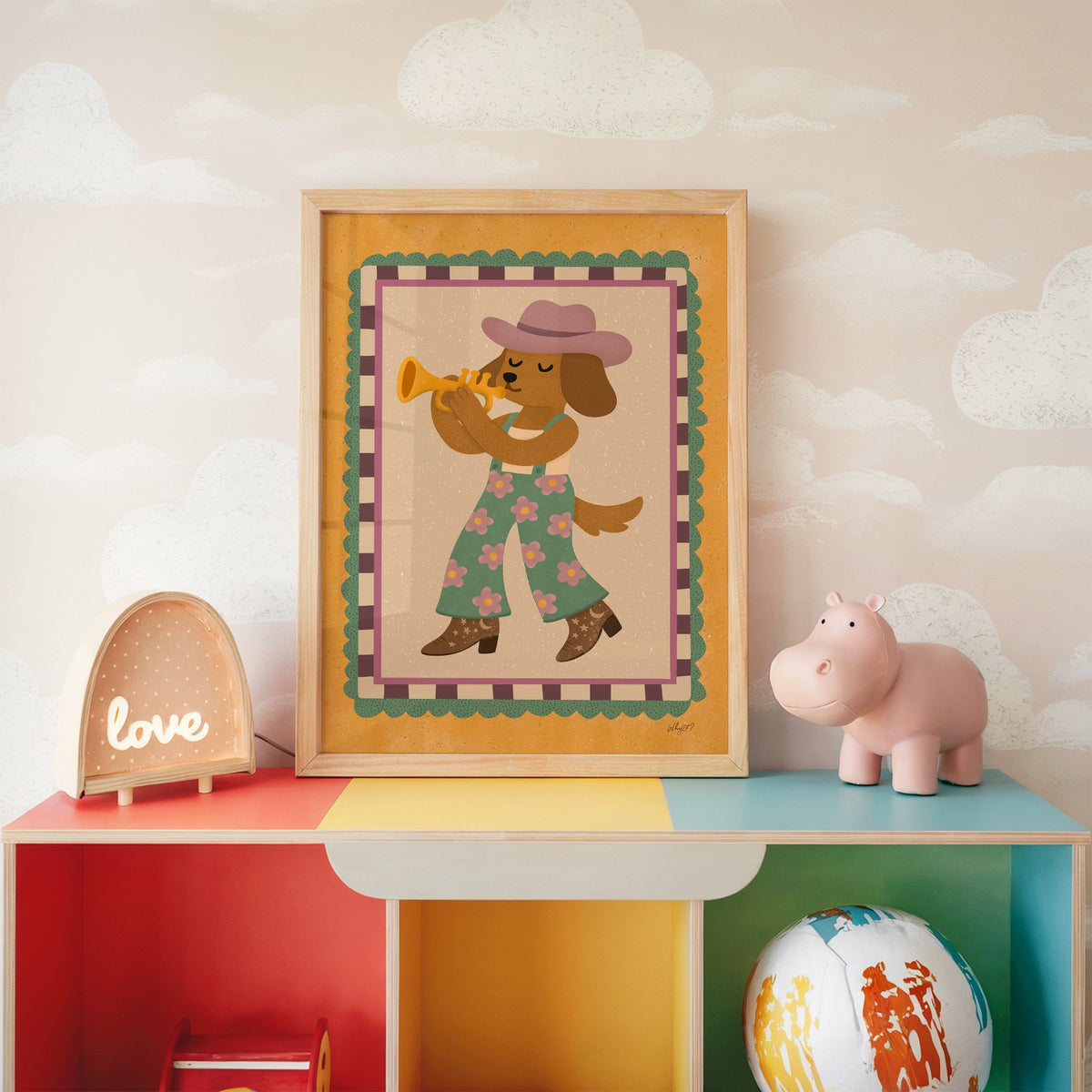 Dog playing trumpet in cowboy hat nursery print in an oak frame on a rainbow toy shelf against cloud wallpaper